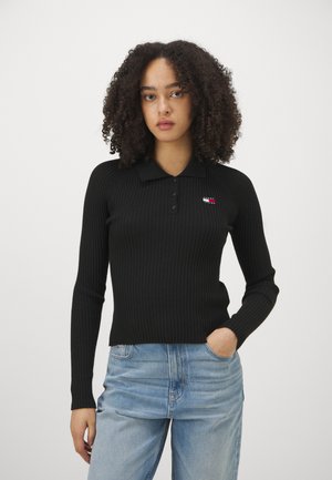 Suéter de polo negro de canalé con mangas largas, que presenta un cuello con solapa y un pequeño parche con el logo. Combinado con jeans de corte relajado en azul claro.