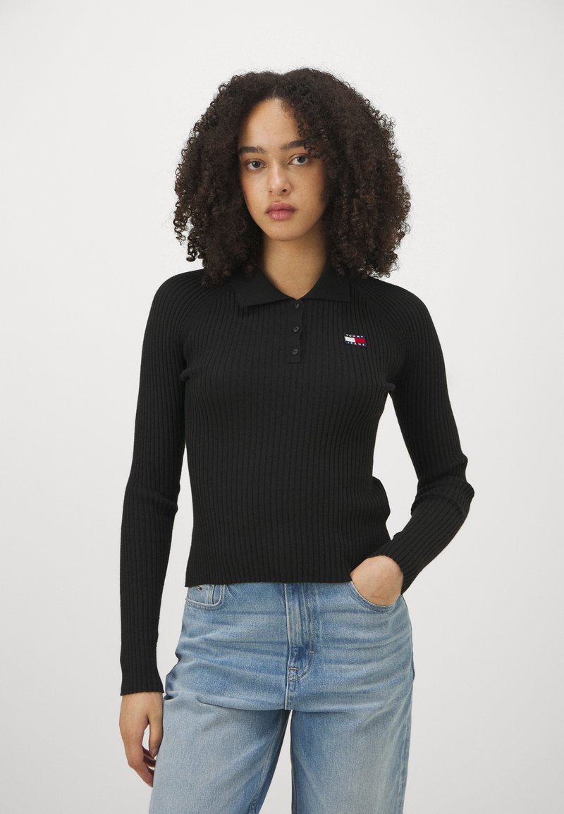 Suéter de polo negro de canalé con mangas largas, que presenta un cuello con solapa y un pequeño parche con el logo. Combinado con jeans de corte relajado en azul claro.