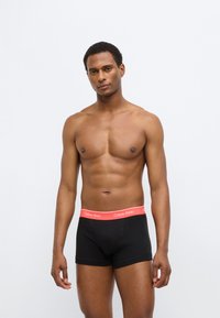 Μαύρα ανδρικά μποξέρ με κόκκινη ζώνη μέσης με το λογότυπο Calvin Klein. Λείο ύφασμα, εφαρμοστή σχεδίαση, μήκος μέχρι το μέσο του μηρού και χωρίς ορατά σχέδια.