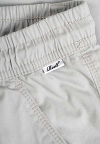 Pantalones gris claro con cintura fruncida, costuras en contraste y una pequeña etiqueta tejida con el nombre de la marca "Reell."