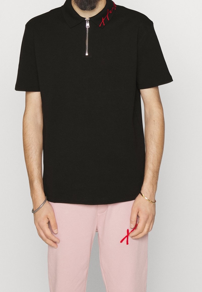 T-shirt polo noir avec un collier zippé, présentant des détails brodés rouges au niveau du col. Porté avec un pantalon rose. Le tissu semble lisse.