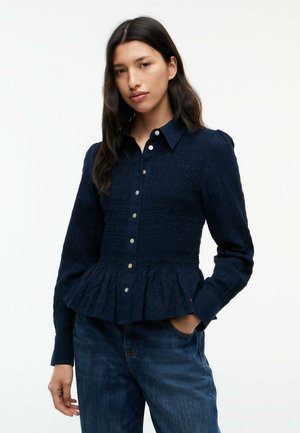 Kvinde med langt mørkt hår iført en mørkeblå tekstureret skjorte med knapper og flæsekant samt blå jeans, stående med den ene hånd i lommen.
