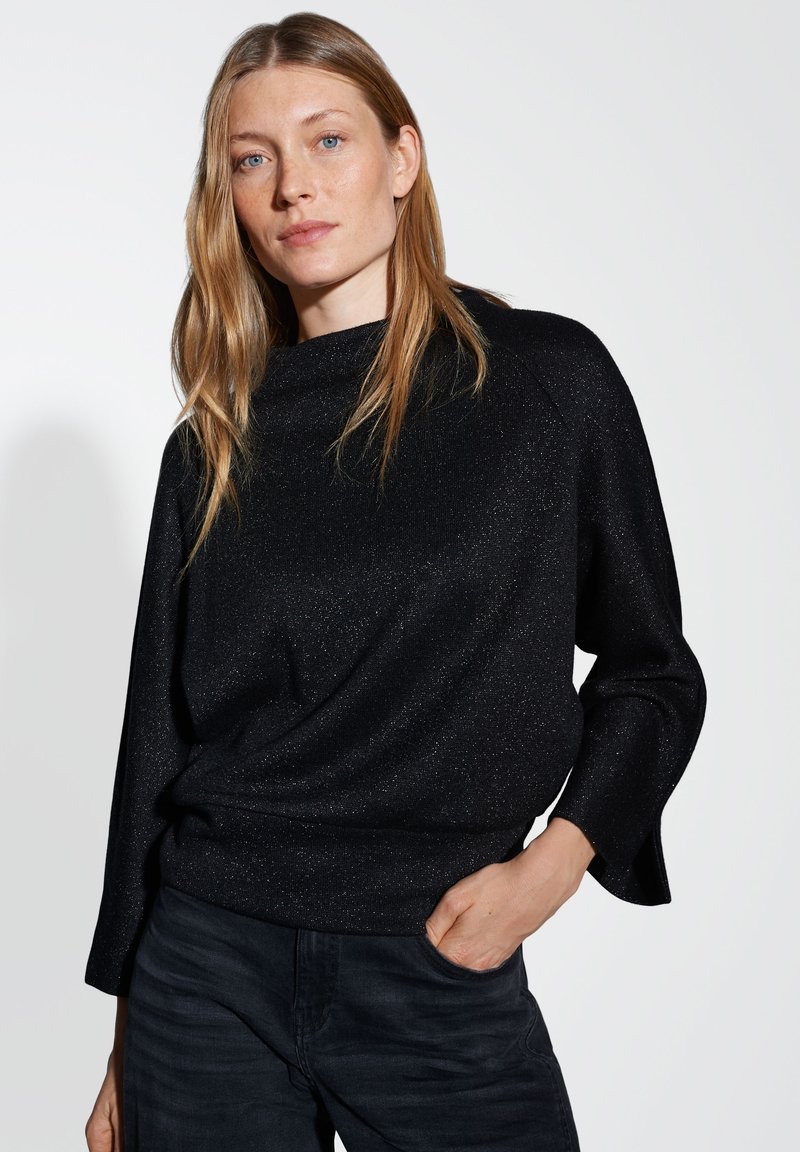 Pull noir scintillant avec un col montant, des manches raglan larges et une coupe décontractée, associé à un jean foncé.