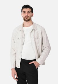Giacca di denim beige chiaro con due tasche sul petto, bottoni argentati e una vestibilità rilassata, abbinata a una t-shirt bianca a girocollo e pantaloni neri.