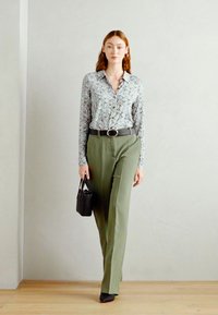 Blůza s květinovým vzorem a knoflíky, zelené kalhoty s vysokým pasem, černý opasek a taška, elegantní černé boty, stojící před světlou zdí.