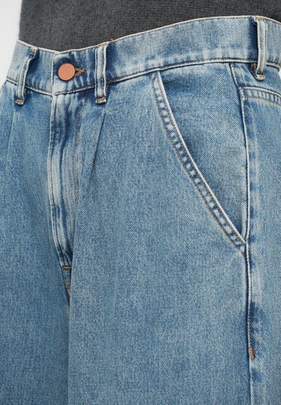 Nærbilde av lys blå denimjeans med folder, frontlommer, beltehemper, og en kobberknapp på en person som har på seg en mørkegrå topp.