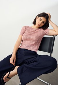 Lyserød ribbet kortærmet skjorte med tekstureret mønster, kombineret med marineblå vide bukser og sorte sandaler, siddende på en metalstol.