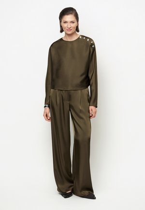 Vrouw draagt een olijfgroene satijnen blouse met lange mouwen en schouderknopen en bijpassende brede broek, staand voor een witte achtergrond.