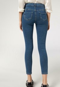 Persona con jeans ajustados de talle alto azules, camisa blanca de manga larga remangada y zapatos negros, de pie con la espalda hacia la cámara.