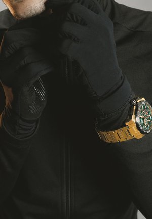 Des gants noirs et une montre dorée sur une personne portant une veste noire, avec une fermeture éclair à l'avant et des accents texturés sur les gants.
