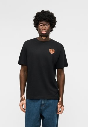 Jeune homme atteint de vitiligo portant un t-shirt noir The North Face et un jean bleu sur un fond clair uni.