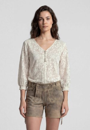 Vrouw draagt een witte bloemrijke kanten blouse met knopen en bruine traditionele leren korte broek staand tegen een effen achtergrond.