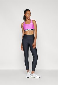 Mujer de pie con leggins negros, sujetador deportivo rosa y zapatillas blancas mirando hacia la derecha contra un fondo liso.