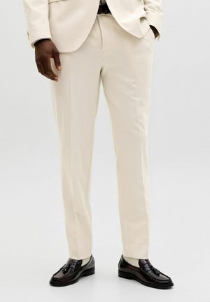 Muškarac obučen u krem hlače po mjeri, krem blejzer, bež čarape i tamnobraon cipele s ukrasnim resama, stoji s rukama u džepovima.