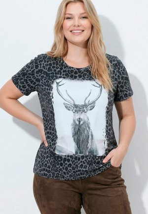 Smilende kvinde med blondt hår iført en sort T-shirt med leopardprint og stort hjortehoved samt brune bukser, stående med hænderne på hofterne.