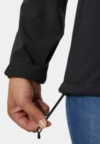 Zwarte jas met een gestructureerde afwerking; close-up van een hand die de koord aanpast aan de onderkant; heeft een ontwerp met omslagmouwen.