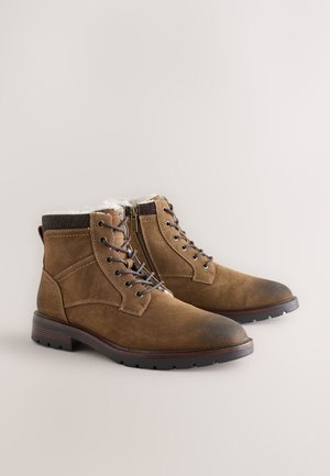 Braune Wildleder-Stiefeletten mit einem weichen weißen Innenfutter, Schnürung vorne, seitlichem Reißverschluss und einer Gummisohle mit strukturierter Oberfläche.