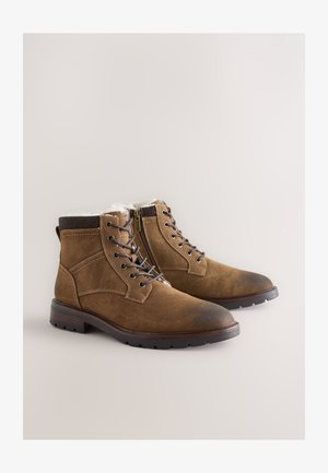 Braune Wildleder-Stiefeletten mit einem weichen weißen Innenfutter, Schnürung vorne, seitlichem Reißverschluss und einer Gummisohle mit strukturierter Oberfläche.