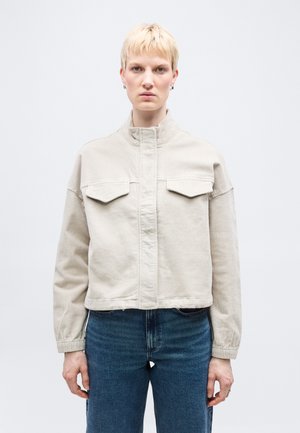 Person mit kurzen blonden Haaren, trägt eine beige Cordjacke mit Vorderentaschen und blaue Jeans, steht vor einem schlichten weißen Hintergrund.