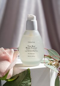 Essenza Riparatrice Vero Rosa URANG in una bottiglia di vetro opaco con un erogatore bianco, etichettata per tutti i tipi di pelle, 50ml, accanto a una rosa rosa e foglie.