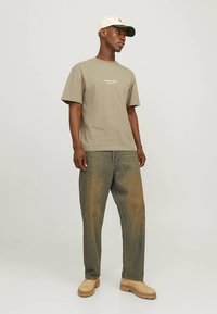 Khaki bavlnené tričko s krátkymi rukávmi a bielym textom, skombinované s voľnými, vyblednutými modrými džínsami a béžovými topánkami, doplnené o svetlú šiltovku.