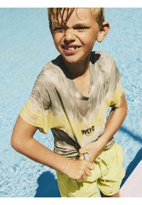 Jongen met kort blond haar staat naast een zwembad, draagt een tie-dye t-shirt in grijs en geel en gele zwembroek, met natte huid.