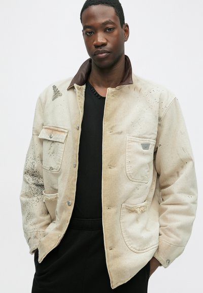Veste en toile de couleur beige clair avec un col marron. Comprend deux poches avant et une texture usée avec des éclaboussures sombres et des bords effilochés.