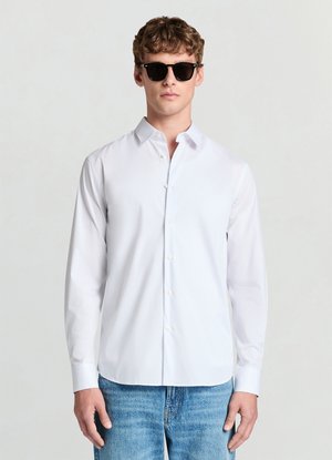 Chemise blanche boutonnée avec col pointu, manches longues et texture lisse, associée à des lunettes de soleil foncées et un jean denim bleu.