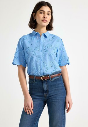 Kvinde iført en lyseblå kortærmet skjorte med blomsterbroderi, stukket ned i højtaljede blå jeans med et brunt bælte.