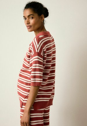 Femme aux cheveux noirs coiffés en chignon, portant un haut en maille à manches courtes rayé rouge et blanc ainsi qu'un pantalon assorti, debout de profil sur un fond uni.