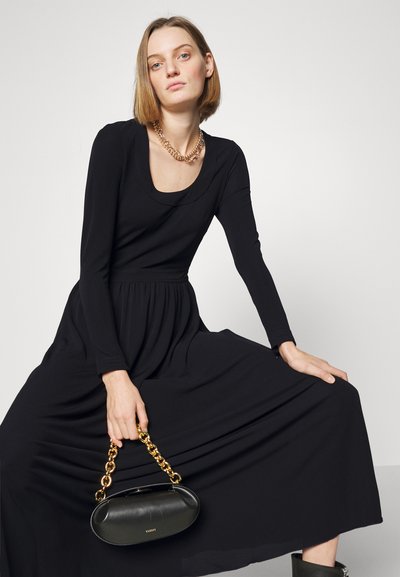 Robe noire à manches longues avec décolleté arrondi, jupe fluide et texture lisse. Le mannequin tient un sac noir arrondi avec une chaîne dorée.