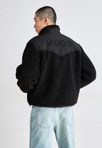 Musta fleece-takki, jossa on kaulusta jäljittelevä yläosa, jossa on musta kangaspaneeli yläselässä, johon on kohokuvioitu "HUGO" paksuilla kirjaimilla.