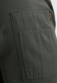 I pantaloni cargo verde scuro presentano una grande tasca cucita sul lato, realizzati in tessuto resistente, con un'etichetta visibile di colore giallo.