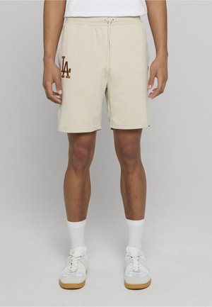 Beige kniehohe Shorts mit Kordelzug und braunem "LA"-Logo, getragen mit weißen Socken und weißen Turnschuhen an einer stehenden Person.