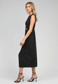 Robe midi sans manches noire avec un col rond, taille froncée et accents latéraux. Comprend des escarpins noirs à lanières. Texture de tissu lisse.