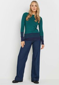 Langärmliges Oberteil in grün und marineblau gestreift mit tailliertem Schnitt und breiten Bündchen, kombiniert mit hoch geschnittenen, locker sitzenden dunkelblauen Jeans.