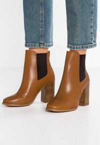 Stivali alla caviglia in pelle marrone con punta affusolata, pannelli laterali elasticizzati e tacco in legno ribattuto. Il design è elegante e presenta una texture liscia.