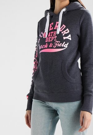 Vrouw draagt een donkergrijze hoodie met witte trekkoorden en roze tekst "Superdry Ath Dept. Track & Field", gecombineerd met lichtblauwe spijkerbroek.