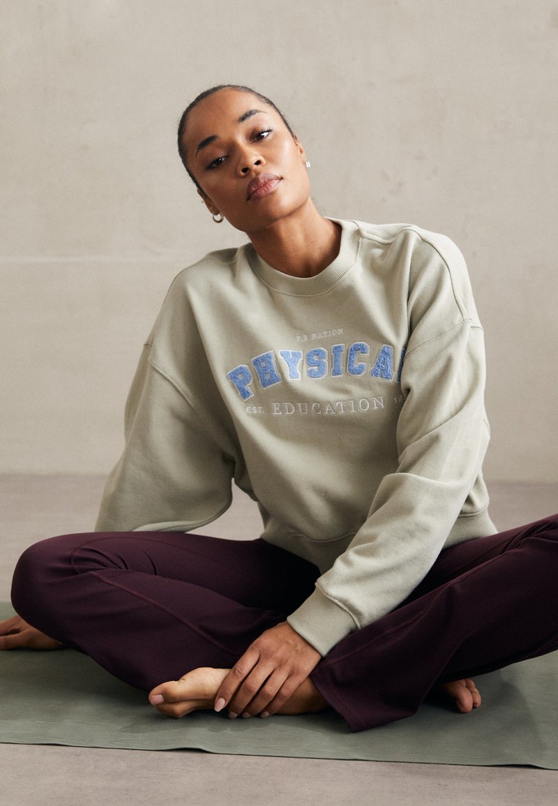 Oversized sage green sweatshirt with "PHYSICAL EDUCATION" embroidered in blue, paired with fitted burgundy pants on a green mat.