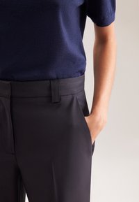 Persoon in een donkerblauwe korte mouwen top, ingerold in een zwarte, op maat gemaakte broek, met de rechterhand in de zak tegen een neutrale achtergrond.