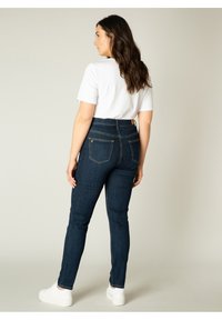 Diepblauwe skinny jeans met een hoge taille, voorzien van drie zakken en subtiele stiksels, gecombineerd met een wit t-shirt en witte sneakers.
