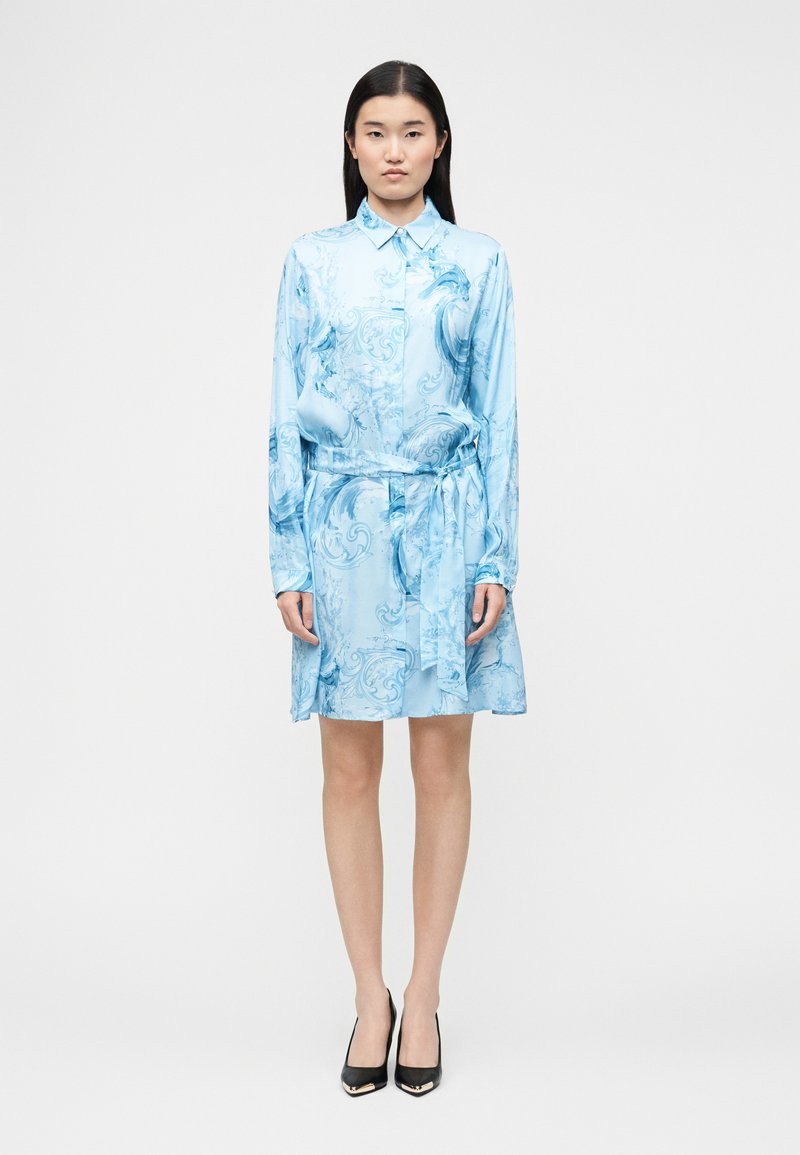 Γυναίκα με μακριά μαύρα μαλλιά που φοράει ένα ανοιχτό μπλε φόρεμα με σχέδιο, μακριά μανίκια και ζώνη στη μέση, καθώς και μαύρα τακούνια, στέκεται όρθια.