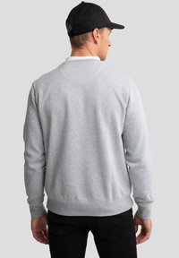 Mand iført en lysegrå langærmet sweatshirt, sorte bukser og en sort baseballkasket, med ryggen mod kameraet på en ensfarvet baggrund.