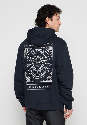 Man die een zwarte hoodie draagt met een witte zon grafiek en tekst "LET THE SUNSHINE INN" en "LYLE & SCOTT" op de rug.