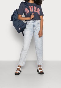 Navy t-shirt with "TOMMY JEANS" in red letters, light blue high-waisted jeans, white sandals with black soles, and a navy backpack.