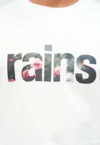 Άσπρο πουκάμισο με τη λέξη «rains» σε μεγάλα γράμματα γεμάτα με ένα λουλουδάτο μοτίβο που περιλαμβάνει ροζ τριαντάφυλλα και πράσινα φύλλα.