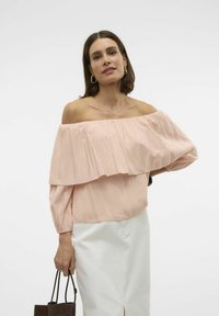 Top rosa scollato spalle, con volants stratificati, maniche lunghe e polsini arricciati; abbinato a una gonna bianca e una borsa a mano marrone scuro.