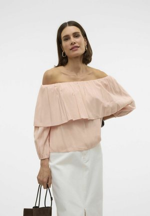 Top rosa scollato spalle, con volants stratificati, maniche lunghe e polsini arricciati; abbinato a una gonna bianca e una borsa a mano marrone scuro.