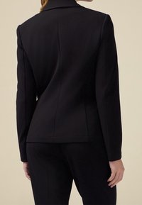 Blazer noir à coupe ajustée, doté d'un col classique, d'un tissu lisse et de détails de couture subtils au dos et à l'ourlet.