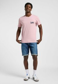 T-shirt in cotone rosa con maniche corte e logo "Lee" blu sul petto, abbinata a pantaloni corti in denim blu e sneakers bianche.